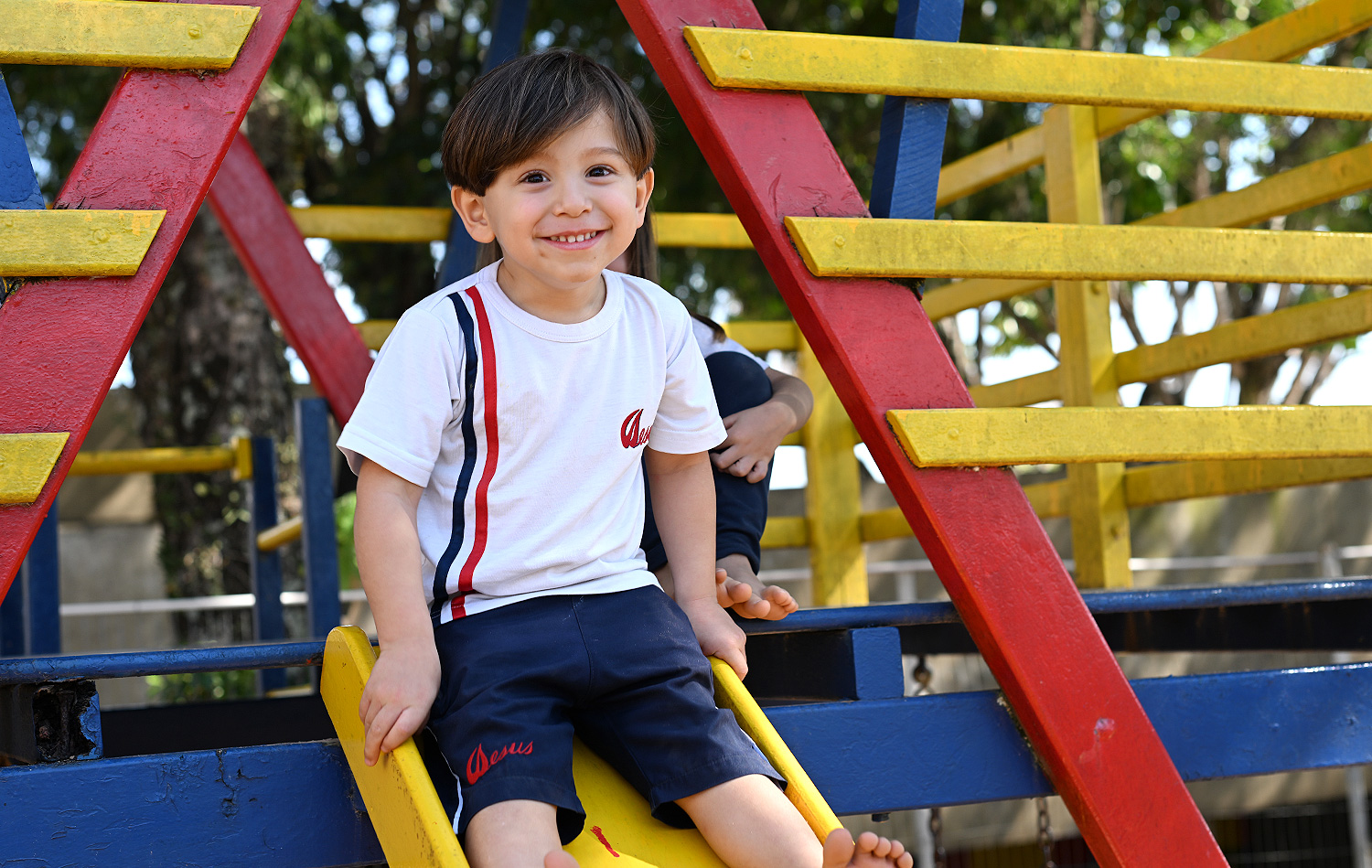 Menino de uniforme branco do colégio sorrindo sentado no alto do escorregador amarelo, entre as estruturas coloridas do parque