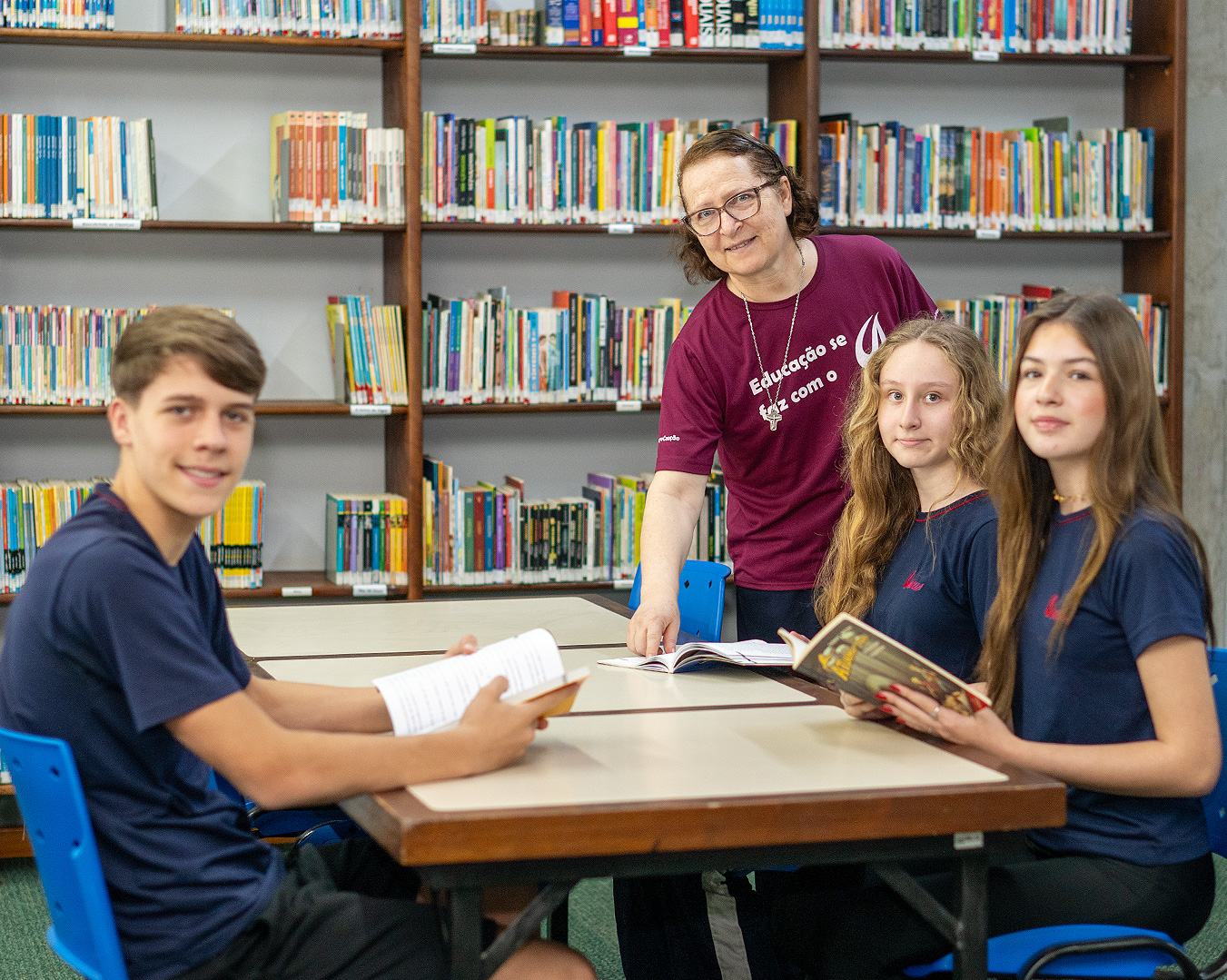 Três estudantes de uniforme azul sentados à mesa da biblioteca com livros abertos, ao lado da professora em pé diante das estantes