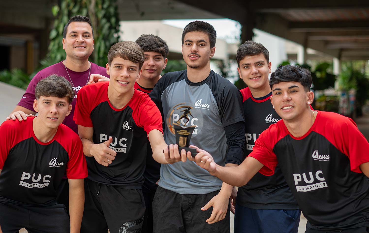 Equipe de estudantes com o professor segurando o troféu de campeões, todos de uniforme esportivo, em área externa do colégio